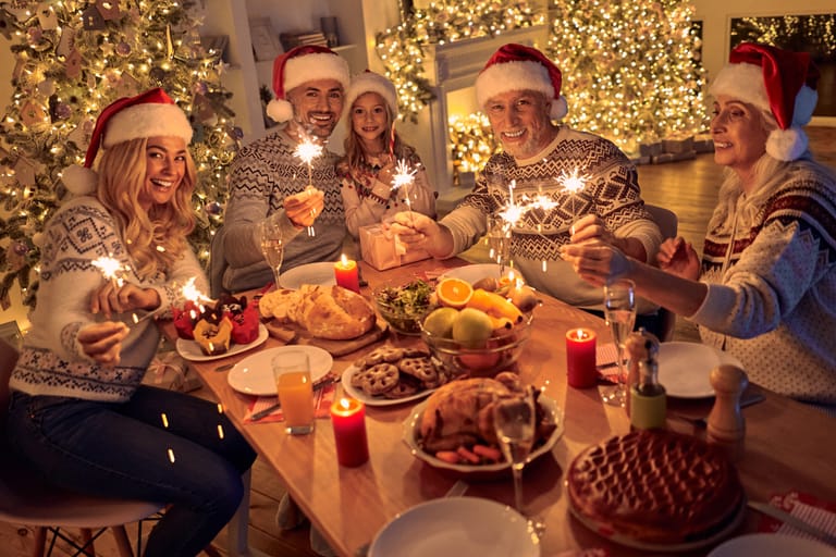 Perhe istuu iltaa joulupöydässä. Kaikilla on jouluiset vaatteet, tonttulakit ja taustalla on jouluiset valot ja ihmisillä on käsissään tähtisädetikut. Kuvituskuva