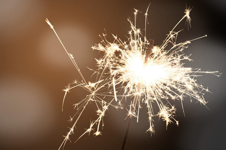Lähikuva palavan tähtisädetikun kipinöistä, kirkas valo ja säkenöivät kipinät erottuvat pehmeästi sumentunutta taustaa vasten.