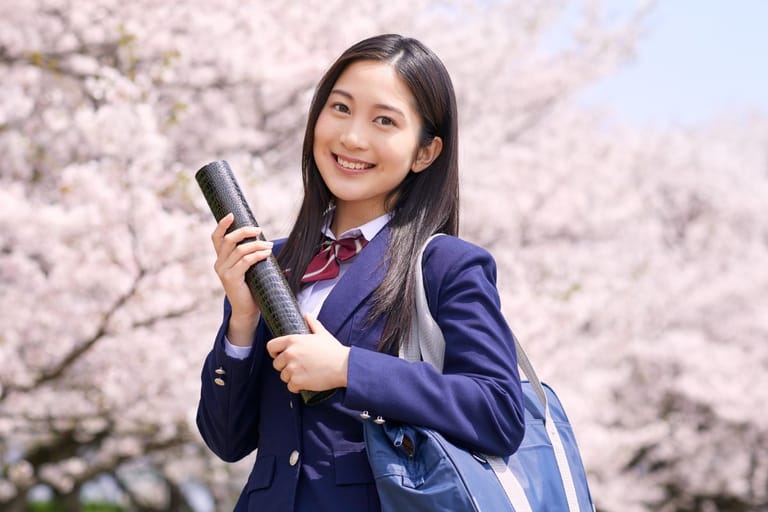 Japanilainen nainen seisoo koulupuvussa kirsikkapuiden keskellä koululaukku mukanaan ja pitelee kädessään jotain mustaa esinettä.