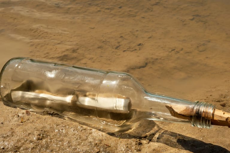 Kuvituskuva. Kuvassa on pullo jonka sisällä on postia, eli pulloposti rantahiekalla päivänvalossa. pullo on läpinäkyvä lasipullo