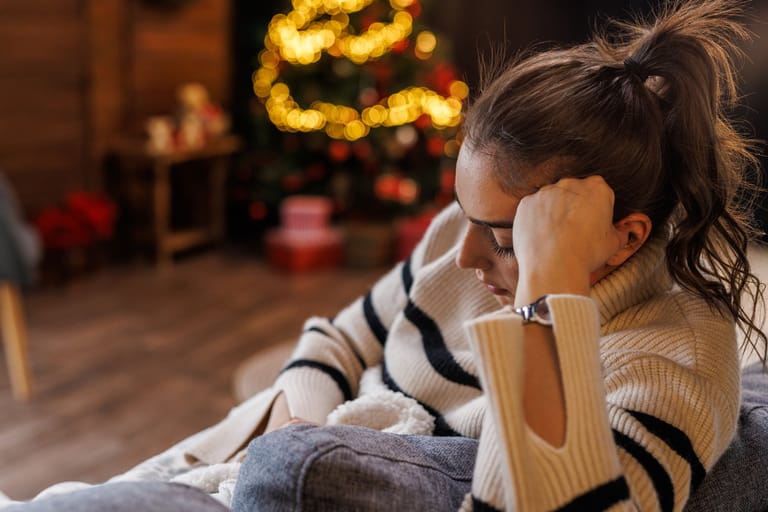 Nainen puhelimella sohvalla. Näyttää surulliselta ja taustalla on joulukuusi kaukaisena haaveena. Kuvituskuva.