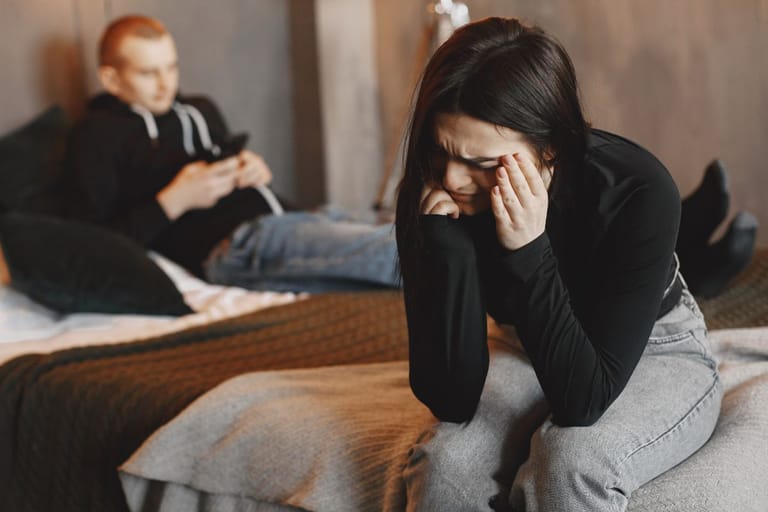 Nainen itkee sängynreunalla ja sängyllä oleva mies ei huomioi. Kuvituskuva
