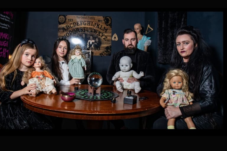 Kuvassa brittiläinen perhe äiti ja isä ja kaksi tytärtä pyöreän pöydän ääressä Addams Family henkisesti pukeutuneina