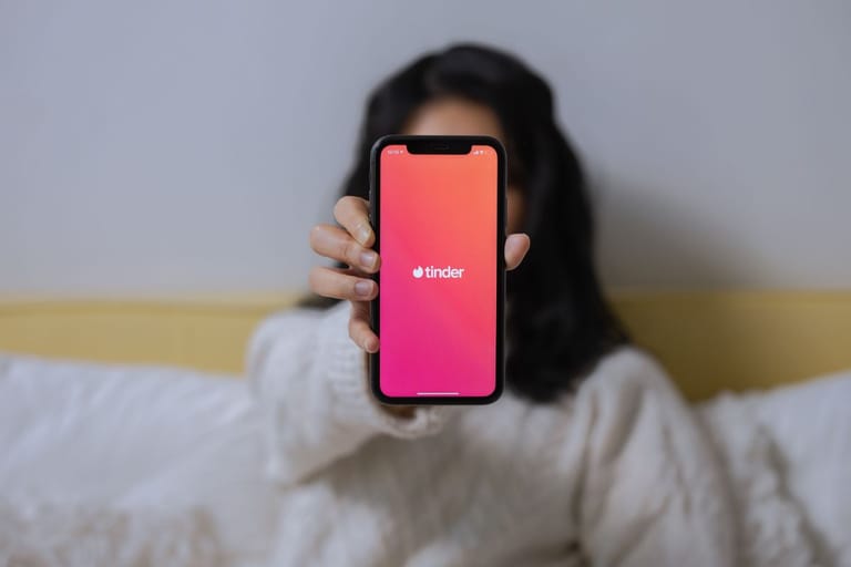 Nainen näyttää Tinderiä sängyllä puhelimensa näytöllä, ja näytöllä on Tinderin logo ja aloituskuva. Nainen on tummatukkainen ja vaaleisiin vaatteisiin pukeutunut.