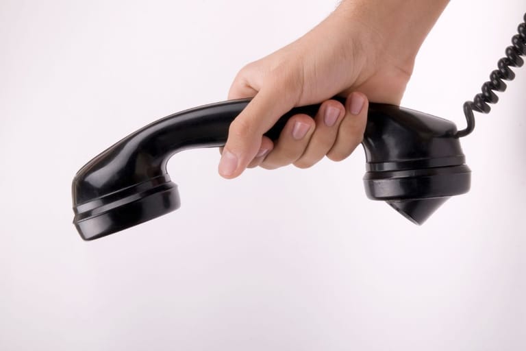 Vanhanaikainen puhelimen luuri ja sitä ollaan laskemassa alas. Mustaa luuria pitää naisen käsi.