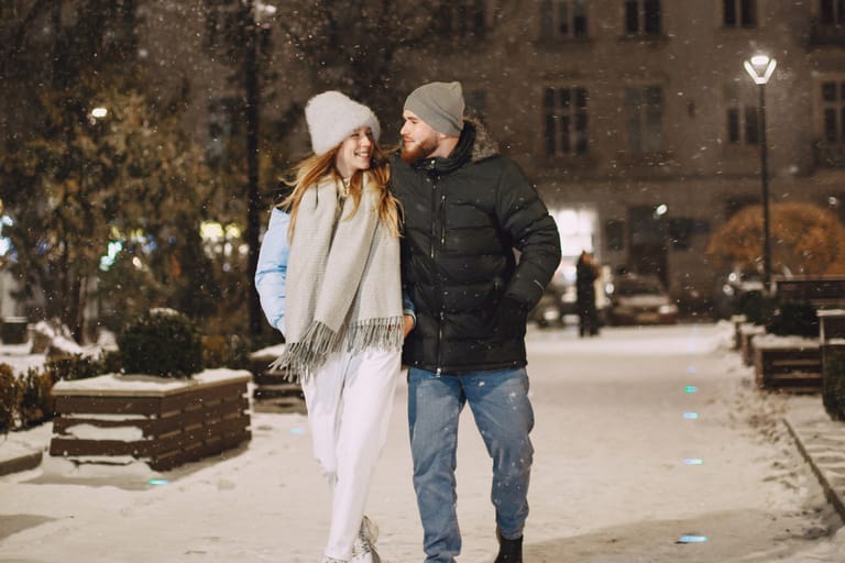 Talvinen iltakatu, jossa nuori pari kävelee vierekkäin käsikynkässä. Molemmilla on talvivaatteet, pipot ja huivit, ja he hymyilevät toisilleen lumisateessa valaistulla kaupunkipolulla.