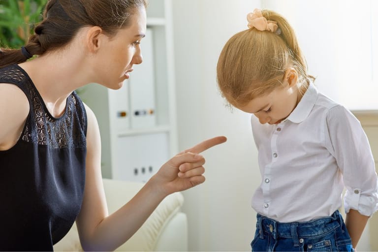 Vanhempi puhuu syyttävästi sormi pystyssä valkoiseen paitaan puetulle lapselle joka näyttää hyvin häpeilevätlä