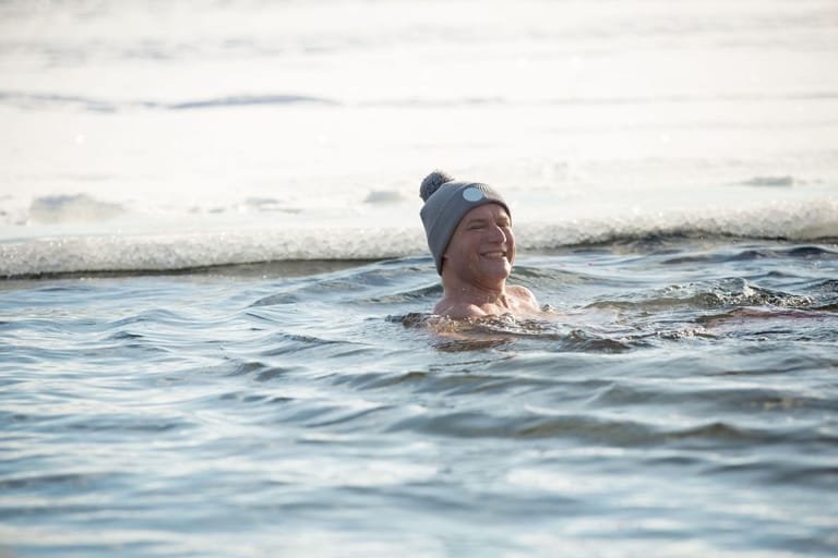 Pipopäinen mies avannossa jäiden keskellä hymyilee kameralle. Avantouintia.