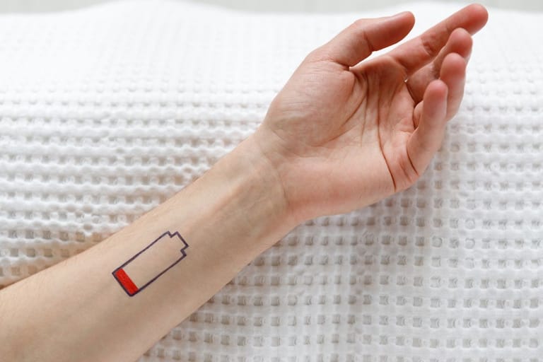 Kuvituskuva uupumuksesta kertovalle artikkelille. Siinä naisen käsi lepää peittoa vasten ja kädessä on tatuointi joka merkkaa patterin loppumista. Kuvituskuva uupumusartikkelille.