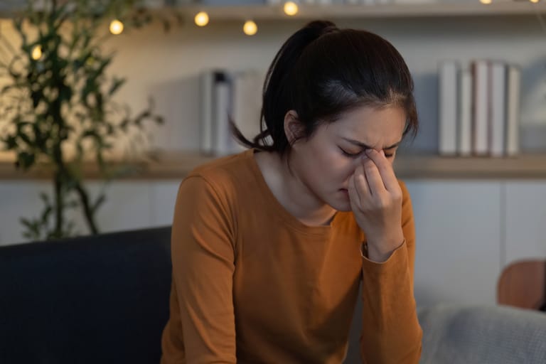 Nuori nainen istuu sisätiloissa ja hieroo silmiään näyttäen väsyneeltä tai stressaantuneelta.