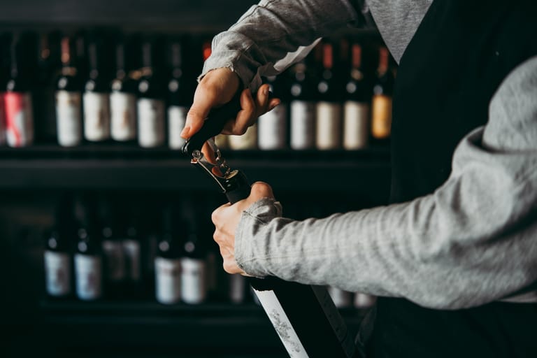 Kuvassa baarimikko avaa viinipulloa. Taustalla näkyy viinipulloja hyllyssä.