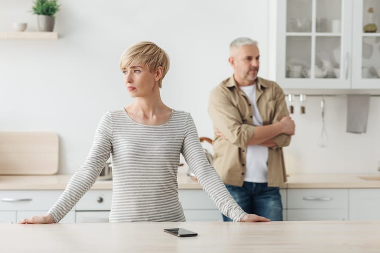 Aikuinen pariskunta seisoo keittiössä toisiinsa selin riidan tai vaikean keskustelun jälkeen. Nainen katsoo poispäin ikkunan suuntaan, kädet pöydällä, ja mies seisoo taustalla kädet ristissä, ilme vakavana. Pöydällä heidän välissään on älypuhelin, joka lisää etäisyyden tunnetta.