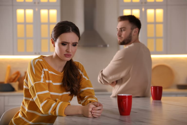 Nainen istuu keittiön pöydän ääressä huolestuneen ja surullisen näköisenä, kädet ristissä edessään. Hänen takanaan mies seisoo kädet ristissä ja katse käännettynä pois. Pöydällä on kaksi punaista mukia. Kuvassa välittyy jännittynyt ja etäinen tunnelma parisuhderiidasta tai vaikeasta keskustelusta.