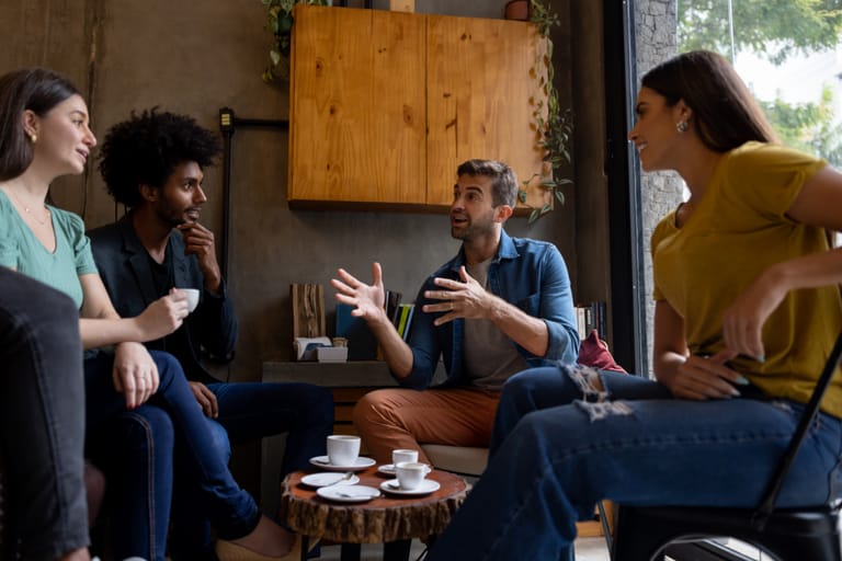 Neljän hengen ystäväporukka istuu kahvilassa pöydän ääressä. Yksi mies puhuu elehtien voimakkaasti käsillään, kun muut kuuntelevat vaihtelevin ilmein. Osa hymyilee, osa näyttää mietteliäältä. Edessä pöydällä on kahvikuppeja ja pieni pöytä kasvien ja hyllyjen reunustamassa tilassa.