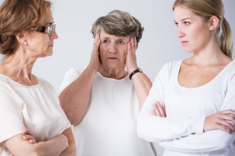 Kolme eri-ikäistä naista seisoo vastakkain jännittyneessä tilanteessa. Keskellä oleva vanhempi nainen pitää käsiään ohimoillaan huolestuneena, kun taas nuorempi ja keski-ikäinen nainen katsovat toisiaan tiukasti kädet ristissä. Kuvassa näkyy sukupolvien välinen ristiriita tai perheriidan hetki.
