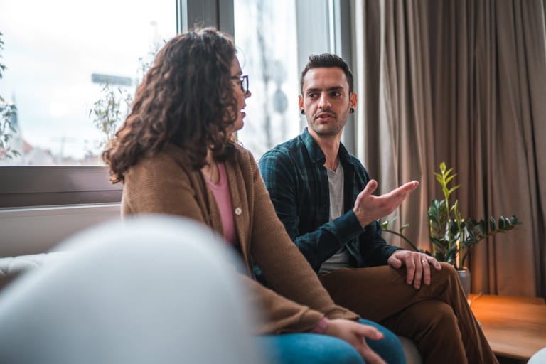 Mies ja nainen istuvat sohvalla keskustelemassa päivänvalossa. Mies puhuu ja elehtii kädellään, nainen kuuntelee keskittyneesti. Tilanne näyttää vakavalta mutta rauhalliselta, taustalla näkyy ikkuna ja viherkasveja.