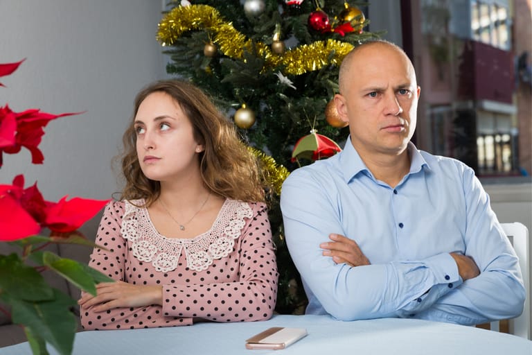 Nuori nainen ja mies istuvat pöydän ääressä ristiriitaisissa tunnelmissa joulukuusen edessä. Molemmat katsovat eri suuntiin, kädet ristissä ja ilmeet jännittyneet. Kuvassa näkyy joulukoristeita ja punaisia kukkia, mutta tunnelma on kireä ja etäinen – kuin riidan jälkeinen hiljaisuus keskellä juhlaa.