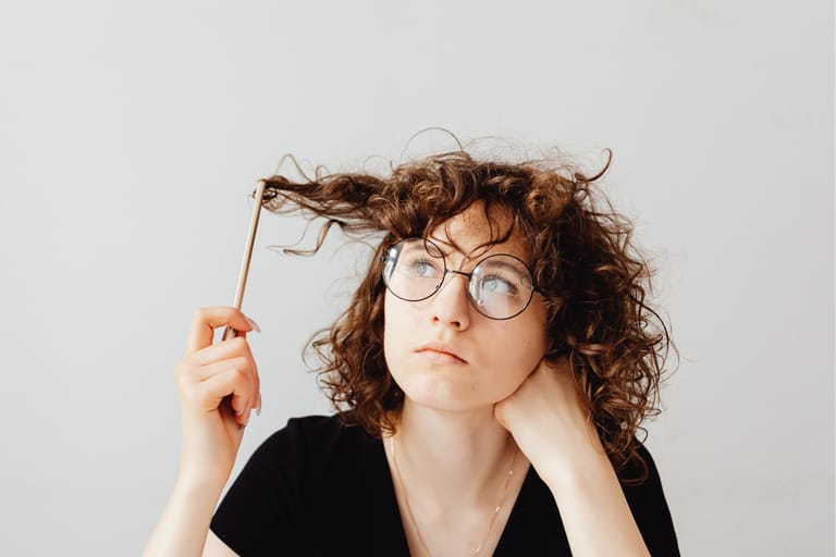 Kuvassa kiharahiuksinen nainen pohtii ja katselee kohti kattoa ja pyörittelee kynällä hiuksiaan.