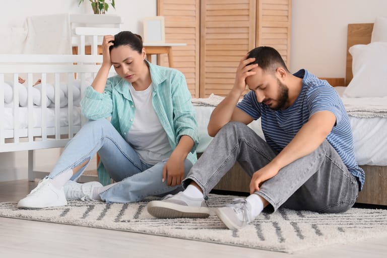 Kuvassa vanhemmat istuvat lastenhuoneen lattialla ja nojailevat uupuneina käsiinsä. Molemmat näyttävät väsyneiltä.