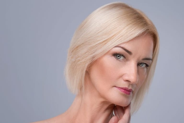 Vaaleahiuksinen, ikääntyvä nainen, joka seisoo vaaleaa taustaa vasten ja jolla on kylmä ilme.