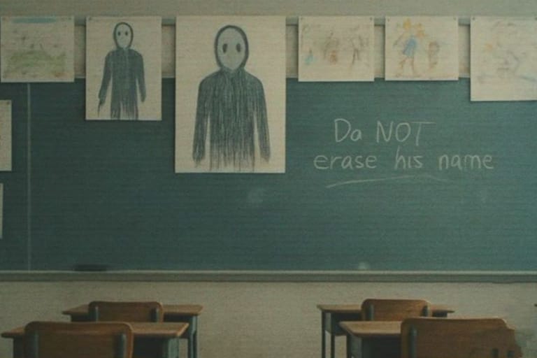 Luokkahuoneen liitutaululle on ripustettu useita lasten piirustuksia, joissa toistuu sama tummaan sävyyn piirretty, suuton ihmishahmo. Liitutaulussa näkyy käsin kirjoitettu teksti: “Do NOT erase his name”. Etualalla on tyhjiä koulupulpetteja.