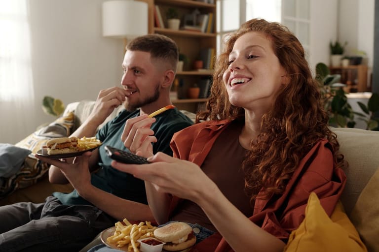 Kaksi aikuista istuu sohvalla olohuoneessa, katsoo televisiota ja syö hampurilaisia ja ranskalaisia. Toinen pitää kädessään kaukosäädintä.