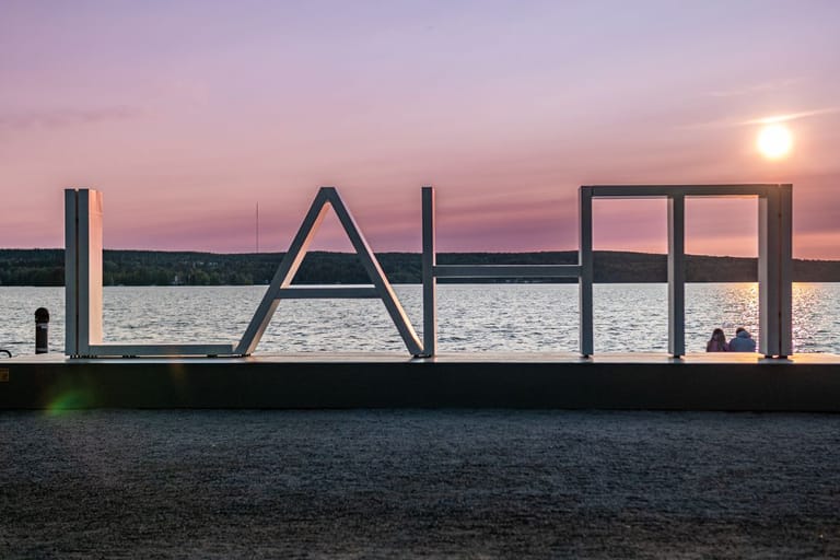 Ilta-auringon valaisema rantamaisema, jossa järven rannalla on suuri metallinen tekstiveistos, jonka kirjaimet muodostavat sanan ”LAHTI”. Taustalla näkyy tyyni järvi ja metsäranta, etualalla hiekkapintainen ranta.