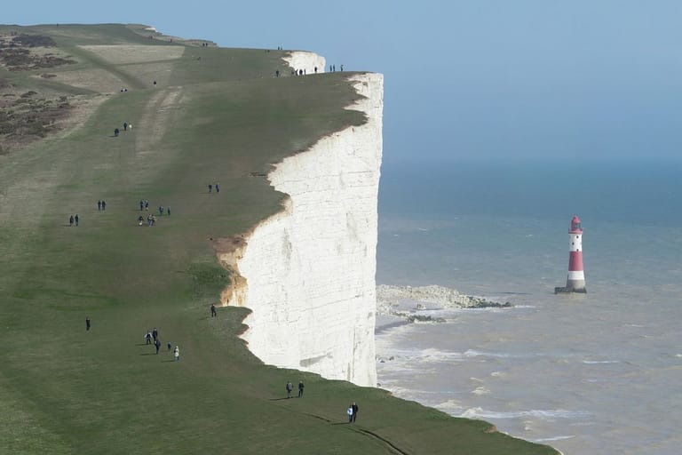 Beachy Headin valkoiset kalkkikivikalliot Etelä-Englannissa. Jyrkänteen reunalla kulkee ihmisiä, ja merellä kallioiden edustalla näkyy punavalkoinen majakka.