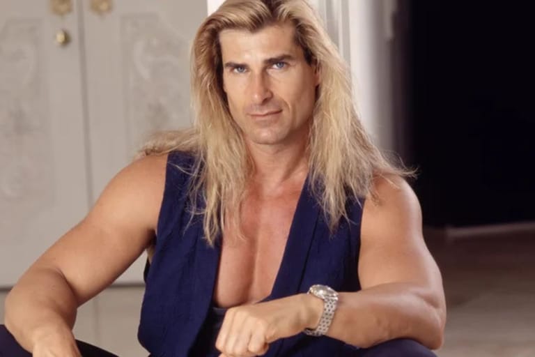 Kuvassa on Fabio Lanzoni, 1990-luvun tunnettu romanssikirjojen kansimalli ja popkulttuuri-ikoni. Hän katsoo suoraan kameraan vakavana, pitkät vaaleat hiukset hartioille laskeutuen ja yllään tummansininen liivi. Kuva henkii tyypillistä 90-luvun glamouria ja Fabion itsevarmaa karismaa.