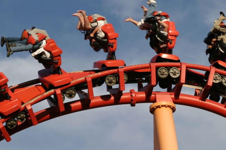 Kuvassa vuoristorata tekee jyrkän silmukan sinistä taivasta vasten. Vaunut ovat kirkkaan oranssit, ja matkustajat roikkuvat ylösalaisin, jalat ilmassa ja hiukset hulmuten vauhdin keskellä. Kuva välittää hurjan liikkeen ja vuoristoradan adrenaliinitunnelman.