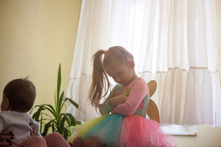 Pieni lapsi seisoo kädet ristissä ja katsoo alaspäin vakavana, kun taustalla aikuinen pitää sylissään vauvaa ikkunan äärellä.