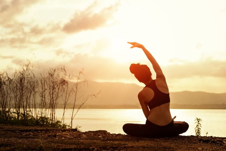 Henkilö istuu maassa järven rannalla ja nostaa toisen käsivarren ylös venytykseen auringonlaskun aikaan.