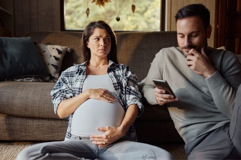 Raskaana oleva nainen istuu sohvalla käsi vatsalla ja katsoo vieressään istuvaa miestä, joka katsoo älypuhelinta.