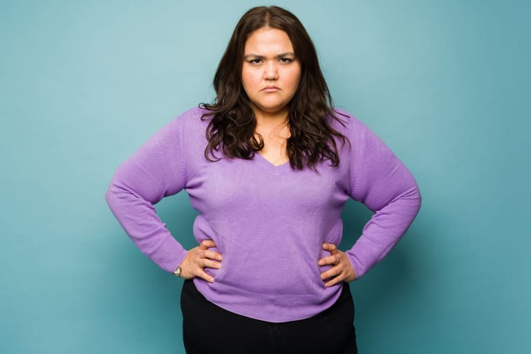 Pluskoon nainen seisoo kädet lanteilla ja katsoo kameraan vaaleaa taustaa vasten.