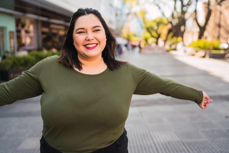 Hymyilevä isokokoinen nainen seisoo ulkona kaupunkimiljöössä kädet levällään. Taustalla näkyy katu ja rakennuksia.