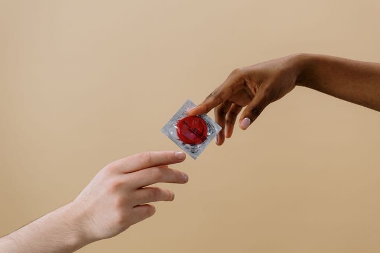 Kaksi kättä ojentaa kondomia toisilleen vaaleanruskeaa taustaa vasten.