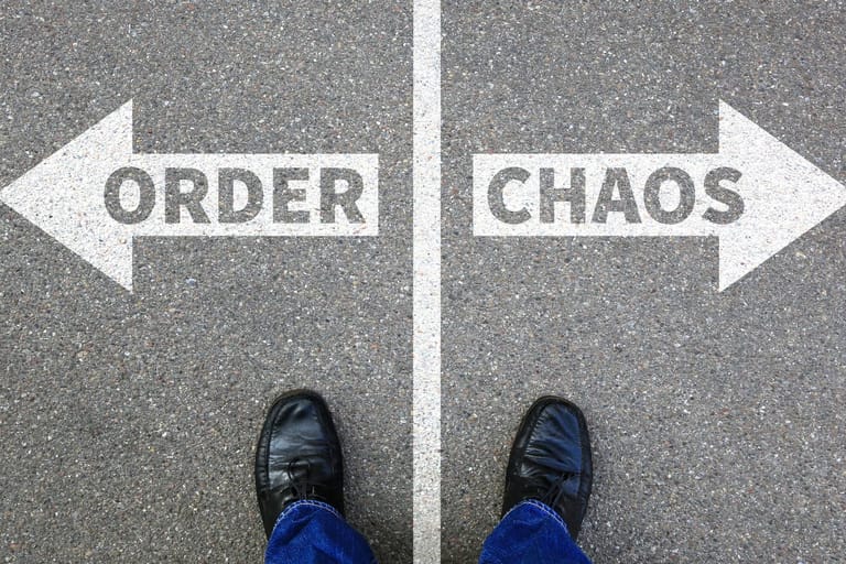 Miehen kengänkärjet osoittavat, että mies seisoo asfaltilla johon on maalattu valkoisella maalilla tienjakaja. Toisella puolella lukee järjestys, toisella kaaos. Kuva symboloi keskitietä.