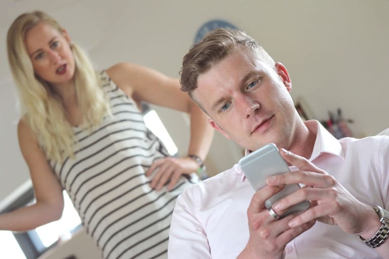 Mies katsoo puhelintaan etualalla, taustalla nainen seisoo käsi lanteilla, ilmapiiri vaikuttaa jännittyneeltä.