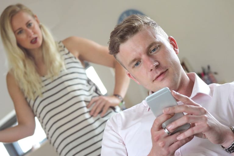 Mies selaa puhelintaan, nainen seisoo taustalla kädet lanteilla epäilevän näköisenä.