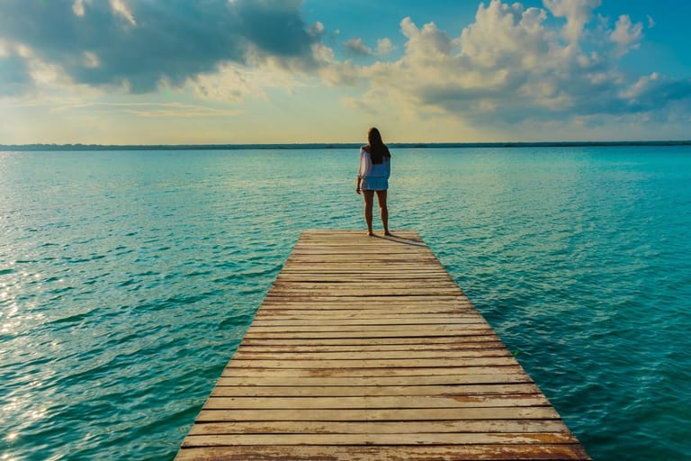 Nainen seisoo unenomaisessa ympäristössä pitkän laiturin päässä ja katselee aavalle ulapalle selkä kuvaajaan päin.