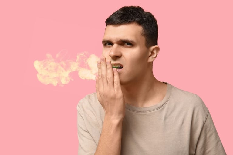 Nuorehko mies vaaleassa paidassa pitää kättään suunsa edessä ja katsoo kameraan. Hänen suustaan leijailee kellertävää savua, joka kuvastaa pahanhajuista hengitystä, vaaleanpunaisella taustalla.