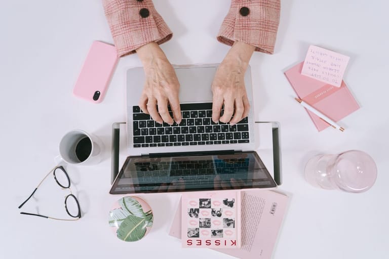 Ylhäältä kuvattu näkymä käsistä kirjoittamassa kannettavalla tietokoneella työpöydällä, ympärillä muistikirjoja ja kahvikuppi.