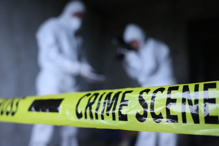 Kaksi valkoiseen suojapukuun pukeutunutta tutkijaa seisoo keltaisten poliisinauhojen takana mustaa taustaa vasten.