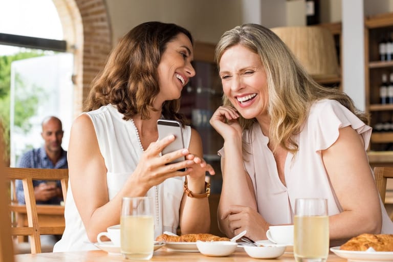 Kaksi naista nauraa yhdessä kahvilassa pöydän ääressä ja katsoo puhelinta. Kuvassa on iloinen ja lämmin tunnelma, joka korostaa yhteyttä ja ystävyyttä – vaikka teknologia onkin läsnä hetkessä.