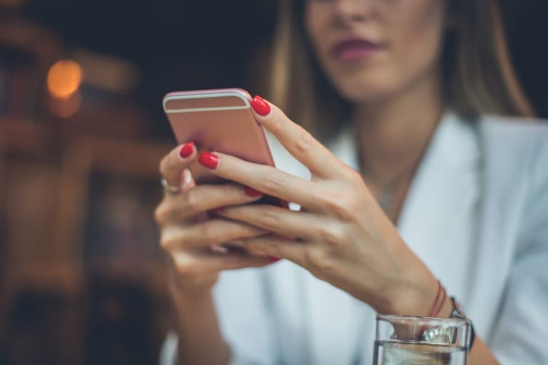 Nainen pitää älypuhelinta käsissään pöydän ääressä, edessään vesilasi.