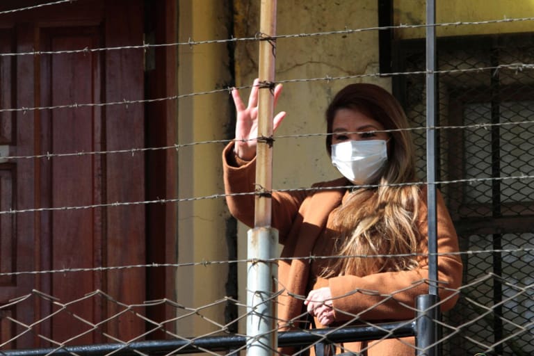 Nainen vilkuttaa piikkilanka-aidan yli alas. Hänellä on kasvoillaan valkoinen maski.