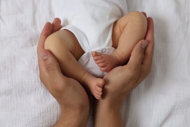 Valkoiseen bodyyn puetun vauvan jalat sekä aikuisen kädet, joka pitää hellästi vauvaa käsiensä välissä valkoista taustaa vasten.