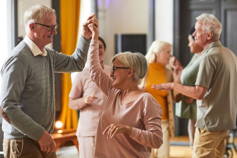 Iäkkäämpi mies ja nainen tanssivat yhdessä sisätilassa, taustalla muita senioreita keskustelemassa ja liikkumassa ryhmätilassa.