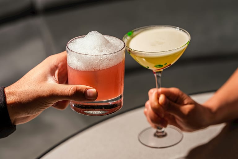 Kaksi kättä kohottaa cocktail-laseja vastakkain. Toisessa lasissa on vaaleanpunainen juoma paksulla vaahdolla matalassa lasissa, toisessa vaalea cocktail jalallisessa lasissa. Tausta on hillitty ja epäterävä, ja kuva välittää intiimin, rennon ja juhlistavan hetken.