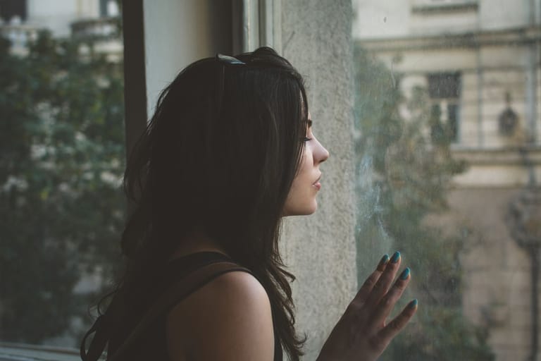 Nainen seisoo ikkunan edessä silmät kiinni ja pitää sormiaan ikkunaa vasten. Ikkunasta näkyy urbaani kaupunkimiljöö.